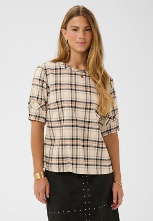 Femme aux longs cheveux blonds porte une blouse beige à carreaux avec des manches retroussées et une jupe noire ornée de petits clous argentés, bijoux aux poignets et aux oreilles.