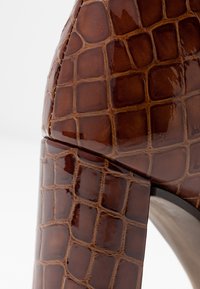 Chaussure à talon haut en cuir verni marron avec un motif texturé crocodile et un talon bloc, présentant un design élégant et une finition brillante.