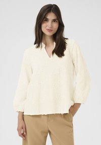 Grietos bluzonai su V formos iškirpte, ilgomis pūstomis rankovėmis ir tekstūruota gėlių siuvinėjimu. Minkšta medžiaga su šiek tiek nusidėvėjusiu kraštu.