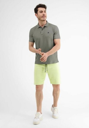 Mann steht und trägt ein olivgrünes Poloshirt, hellgelbe Shorts mit Kordelzug und weiße Turnschuhe vor weißem Hintergrund.