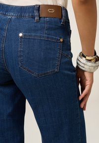 Vaqueros de mezclilla de color azul oscuro con un diseño ajustado, que cuentan con una cinturilla de cuero marrón y un bolsillo trasero con costuras en contraste.