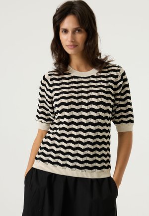 Zwart- en crèmekleurige trui met een zigzagpatroon, korte mouwen en een geribde halslijn, gedragen met een zwarte rok. Gecursiveerde stof.