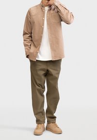 Brązowa koszula sztruksowa na guziki z kieszenią, noszona na białej koszulce; zestawiona z oliwkowozielonymi spodniami i beżowymi tenisówkami.