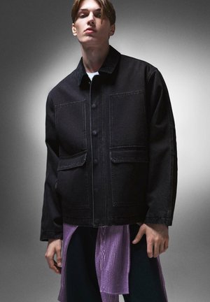 Giacca di jeans nera con ampie tasche anteriori, chiusura con bottoni e cuciture a contrasto. Indossata sopra un indumento testurizzato di colore viola.