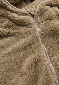 Vaaleanruskea fleece-takki pehmeällä, tekstuuripinnalla, jossa on edessä vetoketju ja korkea kaulus lisälämmön takaamiseksi.