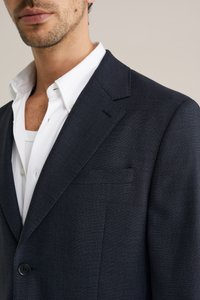 Donkerblauwe blazer van gestructureerde stof, met een enkele knoopsluiting, revers met inkeping en een borstzak, gedragen over een wit overhemd.