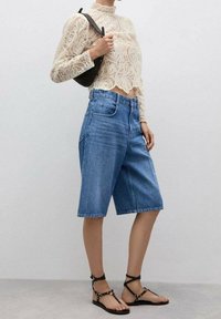 Haut à manches longues en dentelle crème, associé à un short bermuda en denim clair et des sandales à lanières noires avec des accents dorés. Texture lisse et coupe ajustée.