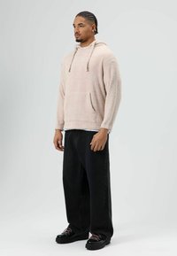 Beige fleece hoodie met een grote voorkantzak en een trekkoord in de capuchon, gecombineerd met loszittende zwarte jeans en zwarte schoenen met roze veters.