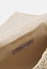 Borsa di paglia intrecciata con rivestimento in tessuto beige. Presenta un'etichetta cucita con la scritta "ANNA FIELD." L'interno ha una finitura liscia e texturizzata.
