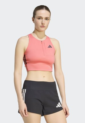 Kvinde iført en koralfarvet ærmeløs crop top med lynlås og sorte atletikshorts med hvide striber og logoer, stående mod en lys baggrund.