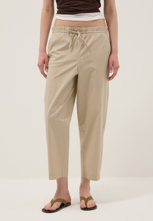 Person wearing beige cropped drawstring pants, brown tank top, white undershirt, and brown flat sandals with studs, standing on plain background.