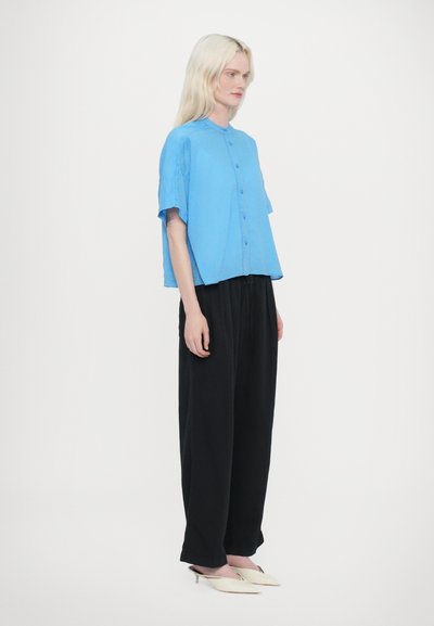 Camicia blu a maniche corte con bottoni abbinata a pantaloni neri larghi. La camicia presenta un colletto tondo e una vestibilità rilassata, creando un look minimalista.