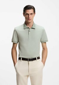Hellgrünes, kurzärmeliges Poloshirt mit strukturiertem Muster, Kragen und zwei schwarzen Knöpfen. Kombiniert mit beigen Hosen und einem dunklen Gürtel.
