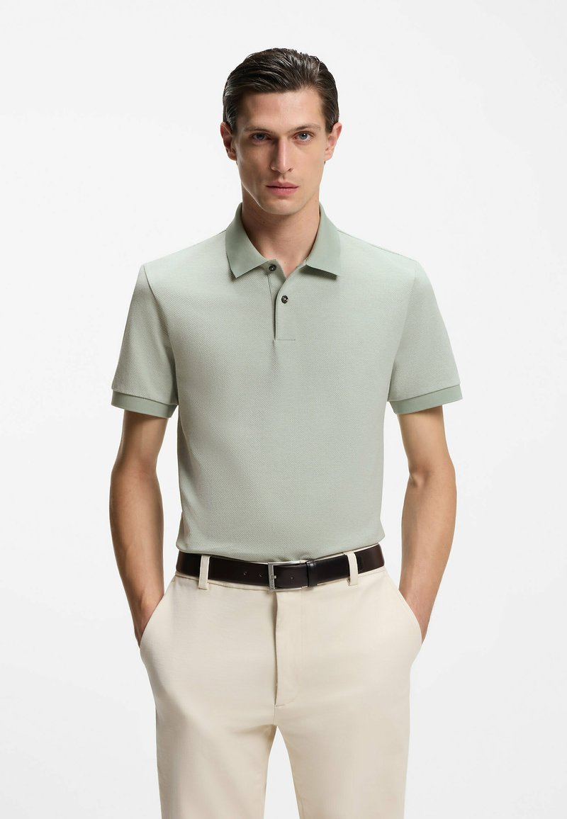 Hellgrünes, kurzärmeliges Poloshirt mit strukturiertem Muster, Kragen und zwei schwarzen Knöpfen. Kombiniert mit beigen Hosen und einem dunklen Gürtel.