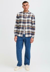 Geruite flanellen shirt in blauw, geel en crème met knopen aan de voorkant; grijs t-shirt daaronder. Blauwe spijkerbroek en witte sneakers maken de look af.