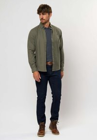 Camicia verde oliva a bottoni, maglione blu navy sotto, jeans blu scuro, stivali marroni con lacci, accessoriata con una cintura marrone e un anello.