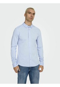 Chemise à manches longues bleu clair avec un col boutonné, fabriquée en tissu doux, associée à un jean foncé. Boutons visibles sur le devant.