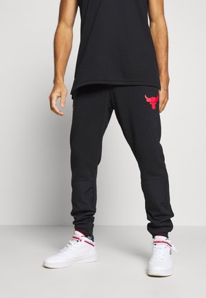 Pantalon de survêtement noir à texture lisse, avec un logo de taureau rouge sur la cuisse gauche, associé à des baskets blanches montantes.