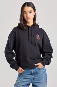 Black hoodie with a front pocket, adjustable drawstring, and a red logo with a sun design on the left chest. Worn with light blue jeans.