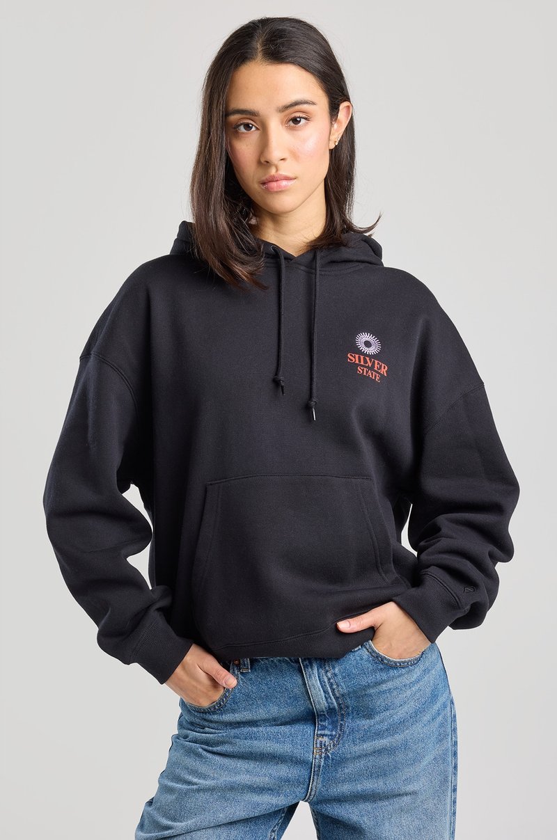 Black hoodie with a front pocket, adjustable drawstring, and a red logo with a sun design on the left chest. Worn with light blue jeans.