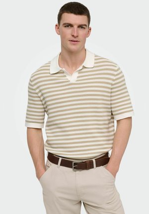 Jeune homme portant un polo à manches courtes en maille rayé beige et blanc, un pantalon beige et une ceinture marron, debout les mains dans les poches sur fond blanc.