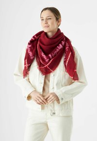 Femme portant une veste en denim écrue et un pantalon, avec une grande écharpe rouge ornée de script rose et de motifs géométriques autour du cou.