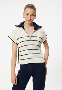 Stickad tröja med dragkedja, i krämfärgad med marinblå ränder, korta ärmar och ståkrage. Texturerad, ribbad material.