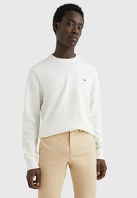 Camicia bianca a maniche lunghe con collo rotondo e polsini a costine, caratterizzata da un piccolo dettaglio del logo sul petto sinistro. Abbinata a pantaloni beige.