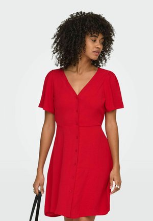 Femme aux cheveux bouclés portant une robe boutonnée rouge à manches courtes, tenant un sac à main noir, regardant vers sa gauche.