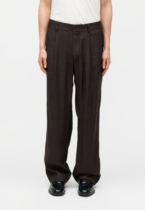 Άτομο με φαρδιά σκούρα καφέ παντελόνια και μαύρα παπούτσια, με τα χέρια χαλαρά στα πλευρά, μπροστά σε απλό φόντο.