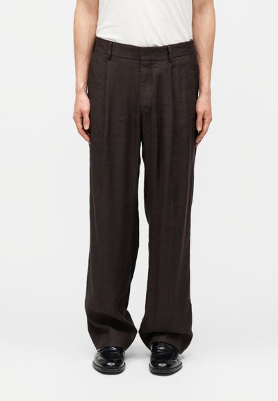 Personne portant un pantalon ample marron foncé et des chaussures noires, les mains détendues le long du corps, sur un fond uni.