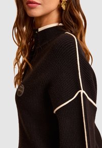 Czarny żebrowany sweter z wysokim dekoltem, kremowymi akcentami na ramionach oraz okrąłym detalem logo na piersi. Teksturowana tkanina.