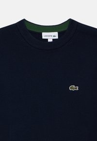 Suéter de punto azul marino con cuello redondo, que presenta un interior de color verde en el cuello y un pequeño logo de cocodrilo bordado en el pecho.