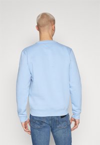Homme avec des cheveux blonds courts portant un sweat-shirt bleu clair et un jean bleu, vu de dos contre un fond blanc uni.