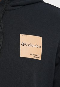 Felpa con cappuccio nera con una toppa rettangolare beige con il logo che presenta il testo "Columbia" e "SPORTSWEAR COMPANY". Tessuto in cotone morbido, design casual.
