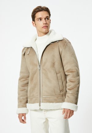 Giacca in suede marrone chiaro con colletto e polsini in finta pelliccia bianca, dotata di chiusura con zip e tasche laterali.
