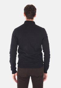 Maglione nero a maglia con colletto alto e polsini a costine, indossato sopra pantaloni marrone scuro, che mostrano una consistenza liscia ed uniforme.