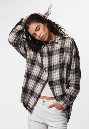 Femme aux cheveux bouclés portant une chemise à carreaux noire et blanche ample sur un top blanc, avec une main dans la poche et l'autre touchant sa tête.