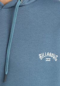 Sudadera azul de algodón con capucha ajustable, logo bordado de "Billabong" en blanco y una textura suave sobre un fondo sólido.