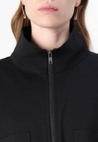 Čierna zipová bunda s vysokým golierom, vyrobená z hladkého materiálu. Obsahuje strieborný zips a dve horizontálne vrecká na hrudi.