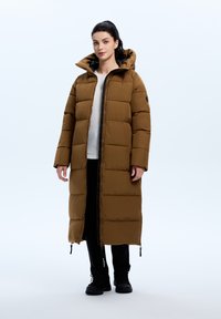 Lange, bruine pufferjas met een capuchon, met een rits aan de voorkant en horizontale stiksels. Draagt een witte top en zwarte broek en schoenen.