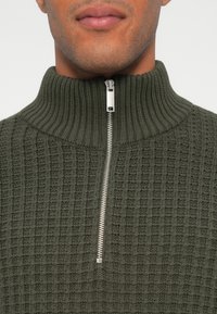 Pulover verde măsliniu cu fermoar la jumătate, cu un model de tricot texturat, guler ribbed și fermoar argintiu. Potrivit pentru suprapuneri sau pentru purtarea în stil casual.