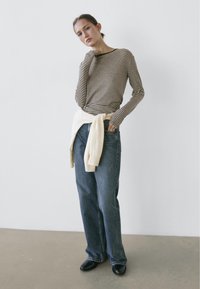 Langarmshirt mit schwarzen und beigen Streifen, kombiniert mit locker sitzenden blauen Jeans. Ein cremefarbener Pullover um die Taille gebunden. Schwarze Schuhe.