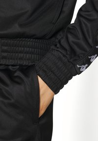 Main dans la poche d'un pantalon de sport noir avec ceinture élastique et manche de veste noire assortie ornée d'une bande répétée avec un logo blanc.