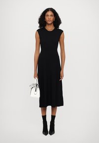 Vestido preto canelado sem mangas com um corpete justo e uma saia rodada, combinado com uma bolsa branca com detalhes pretos e calçado de salto alto.