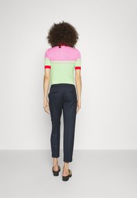 Personne aux cheveux bouclés se tient face à un mur blanc, portant un haut à manches courtes rose et vert, un pantalon bleu marine et des sandales compensées noires.