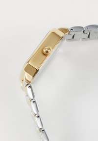 Zlaté a stříbrné obdélníkové hodinky s kovovým páskem, vybavené texturovanou korunou a hladkými okraji pouzdra. Leštěný povrch po celém obvodu.