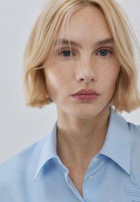 Cămașă albastru deschis cu guler și nasturi, având o textură netedă și detalii subtile de design, purtată de o persoană cu păr blond.