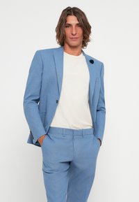 Blazer ajusté bleu clair et pantalon assorti. Le blazer présente un col à revers et une fermeture à bouton unique. Texture douce avec un motif minimal.