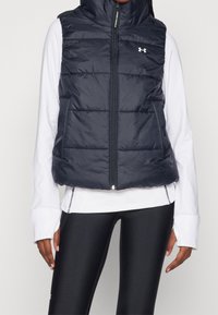 Marineblaues Steppweste mit hohen Kragen, Reißverschluss und horizontalem Steppmuster; über einem weißen Langarmshirt und schwarzen Leggings getragen.